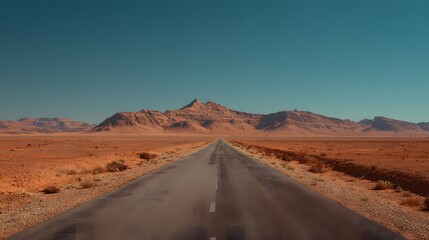 A long straight road cuts through a vast desert landscape towards imposing orange mountains under a clear blue sky