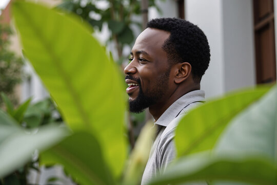 happy and relaxed Black man with a beard smiles warmly while seen through green leaves, enjoying a peaceful moment outdoors in a garden or park with a positive expression