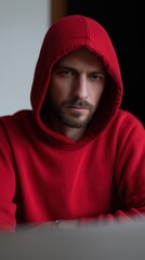 A white hacker in a red hoodie is working on a laptop in a dimly lit room with a light wall and a dark door.