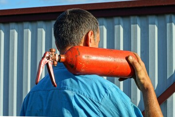 one red old rusty carbon dioxide in a cylinder under pressure dangerous industrial iron with a handle fire extinguisher in the hand of a man on the shoulder in a blue shirt near a gray fence