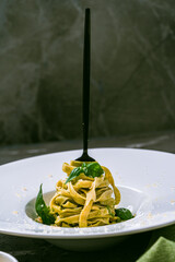 Fituccine al pesto pasta with basil and pine nuts on a white plate with a black fork on a dark gray...