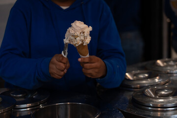 A hand in a blue hoodie scoops velvety ice cream into a crispy waffle cone inside a modern parlor,...