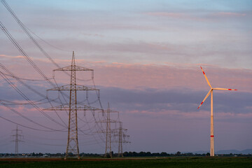 Power transmission lines and a wind turbine dominate the horizon above green fields under a...
