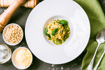 Italian cuisine. Fituccini al pesto on a white plate with a green napkin and cutlery with basil, pine nuts and Parmesan cheese. View from above.