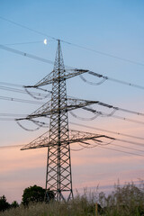 Fototapeta premium A high-voltage transmission pylon stands silhouetted against a vibrant sunset sky with the moon peeking through clouds, blending industrial structure and natural beauty