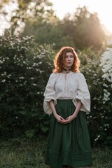 Young pretty caucasian woman in romantic outfit walking in blooming spring garden. Artistic outdoor photo of beautiful red-haired girl posing in fragrant lilac blossom.