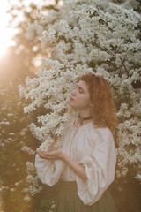 Young pretty caucasian woman in romantic outfit walking in blooming spring garden. Artistic outdoor photo of beautiful red-haired girl posing in fragrant lilac blossom.