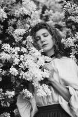 Young pretty caucasian woman in romantic outfit walking in blooming spring garden. Artistic outdoor photo of beautiful red-haired girl posing in fragrant lilac blossom.