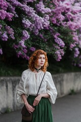 Young pretty caucasian woman in romantic outfit walking in blooming spring garden. Artistic outdoor photo of beautiful red-haired girl posing in fragrant lilac blossom.
