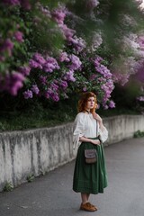 Young pretty caucasian woman in romantic outfit walking in blooming spring garden. Artistic outdoor photo of beautiful red-haired girl posing in fragrant lilac blossom.