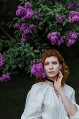 Young pretty caucasian woman in romantic outfit walking in blooming spring garden. Artistic outdoor photo of beautiful red-haired girl posing in fragrant lilac blossom.