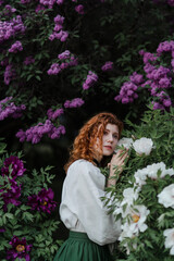 Young pretty caucasian woman in romantic outfit walking in blooming spring garden. Artistic outdoor photo of beautiful red-haired girl posing in fragrant lilac blossom.