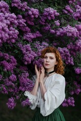 Young pretty caucasian woman in romantic outfit walking in blooming spring garden. Artistic outdoor photo of beautiful red-haired girl posing in fragrant lilac blossom.
