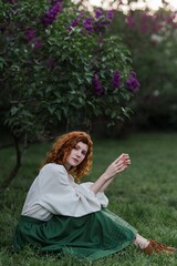 Young pretty caucasian woman in romantic outfit walking in blooming spring garden. Artistic outdoor photo of beautiful red-haired girl posing in fragrant lilac blossom.