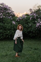 Young pretty caucasian woman in romantic outfit walking in blooming spring garden. Artistic outdoor photo of beautiful red-haired girl posing in fragrant lilac blossom.