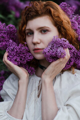 Young pretty caucasian woman in romantic outfit walking in blooming spring garden. Artistic outdoor photo of beautiful red-haired girl posing in fragrant lilac blossom.