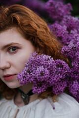Young pretty caucasian woman in romantic outfit walking in blooming spring garden. Artistic outdoor photo of beautiful red-haired girl posing in fragrant lilac blossom.