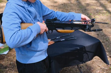 Teenager in blue hoodie loading black sport crossbow at shooting range in forest. Active...