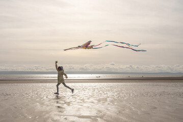 Child Flying Kite on Reflective Welsh Beach. A child runs across a glistening wet beach, joyfully flying a large, colorful kite against a bright, cloudy sky and vast ocean and sea horizon.