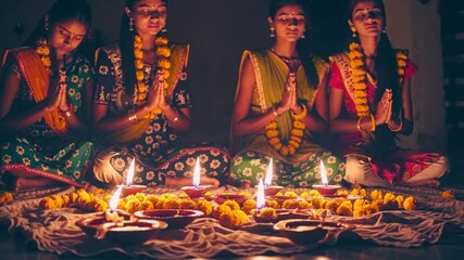 Indian women in traditional saris praying together with joined hands near lit diyas and yellow flower garlands during diwali festival celebration - Powered by Adobe