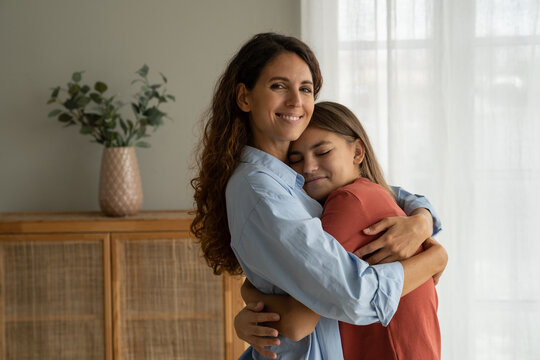 Young loving mother hugging cuddling with teen girl daughter at home and smiling at camera, expressing unconditional love. Mother-daughter friendship, happy motherhood, parenting of adolescent