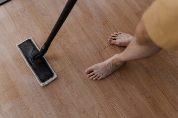 High-angle view of barefoot male cleaning hardwood floor with steam mop during house cleaning,...