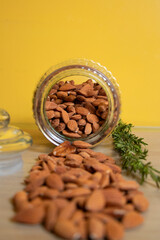 Almonds spilling from glass jar on wooden surface with rosemary and yellow background
