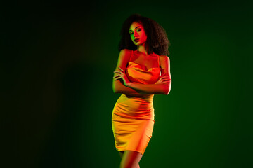Beautiful young woman in elegant orange dress with vibrant green and red lighting on gradient...