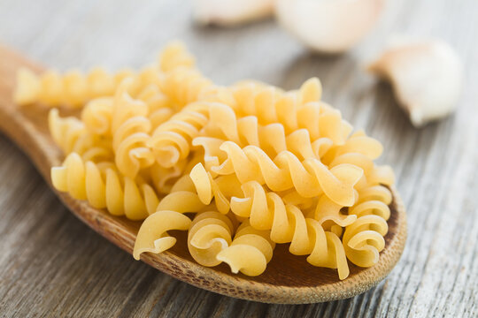 Raw fusilli or rotini pasta on wooden spoon, with garlic cloves in the back (Selective Focus, Focus in the middle of the image)