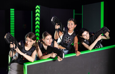 Young women in laser tag studio play safe military tactical game, hide behind barrier, protect...