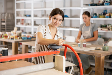 During classes in creative workshop, girl interacts with machine clay press roller to prepare...