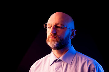 A man with a beard and glasses is standing in front of a black background. Male model in light shirt, bald head illuminated with orange and blue light. Office worker studio shot.