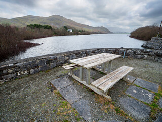 A wooden picnic table sits on a gravel area next to a lake. The table is empty and the surrounding...