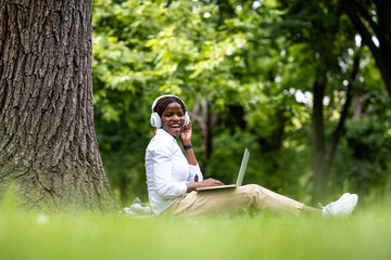 Cheerful Afro girl with headset working on laptop computer in city park. Enjoying the nature.