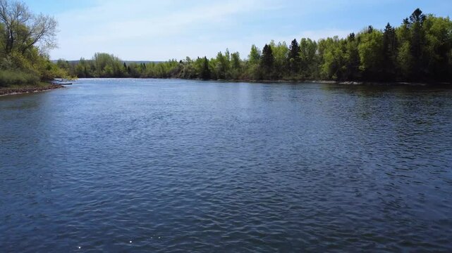 Scenic drone footage of the shimmering Mitis River flowing through a tranquil green countryside with a distant church steeple, Price, Quebec, Canada