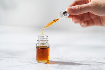 A hand holding a dropper above a small glass bottle filled with amber liquid. The background is a light marble surface, creating a clean and modern aesthetic.
