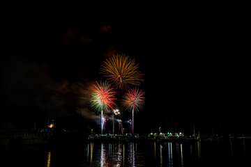 Colorful fireworks exploding over a marina at night, reflected in the calm harbor water with yachts and masts silhouetted under a starless sky with copy space