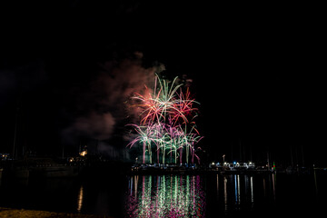 Colorful fireworks exploding over a marina at night, reflected in the calm harbor water with yachts...