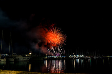 Colorful fireworks exploding over a marina at night, reflected in the calm harbor water with yachts...