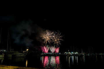 Colorful fireworks exploding over a marina at night, reflected in the calm harbor water with yachts and masts silhouetted under a starless sky with copy space