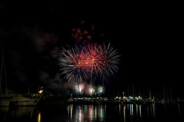 Colorful fireworks exploding over a marina at night, reflected in the calm harbor water with yachts and masts silhouetted under a starless sky with copy space