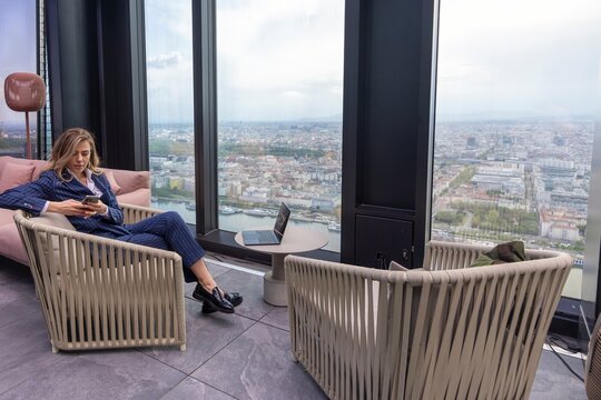 A stylishly dressed woman is seated comfortably in an elegantly designed lounge area, savoring a breathtaking panoramic view of the bustling city skyline from the comfort of a highrise building
