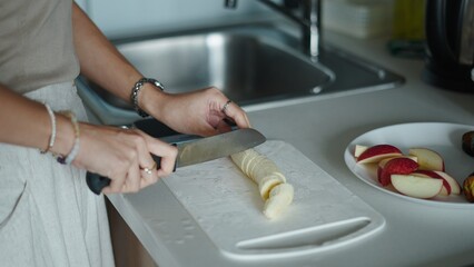 Woman slicing banana on cutting board, preparing healthy breakfast with fresh fruit in modern...