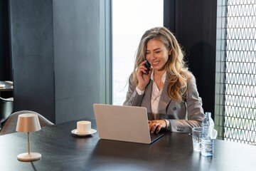 A dedicated professional woman on a phone call while working on her laptop in a modern office,...