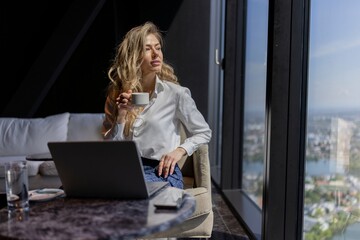 A dedicated professional is working at her modern workspace, sipping coffee and engaging with her laptop, enjoying the stunning city views that boost her creativity and productivity