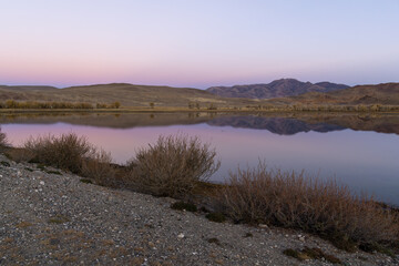 Serene landscape at dusk featuring calm water reflecting mountains and soft pastel sky, surrounded...