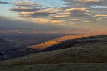 Expansive landscape showcasing rolling hills under a dramatic sky filled with clouds, illuminated by warm sunlight, creating a serene and tranquil atmosphere in nature's beauty