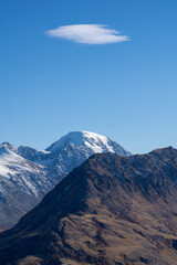 Majestic mountain range with snow-capped peaks under a clear blue sky, showcasing rugged terrain and a unique cloud formation above the highest summit, evoking natural beauty