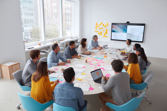 Diverse team collaborating in a modern office. Brainstorming session around a table with tech, notes, and digital display. Perfect for teamwork, innovation, and growth.
