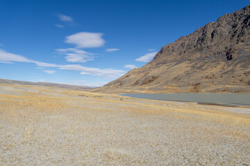 Expansive dry landscape with golden grass, rocky mountains in the background, and a calm lake...